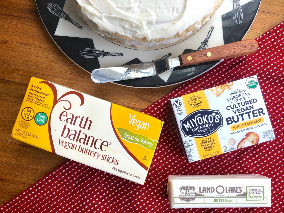 Containers of Earth Balance, Miyoko's, and Lane O Lakes butters on a wooden table, frosted valilla cake alongside.